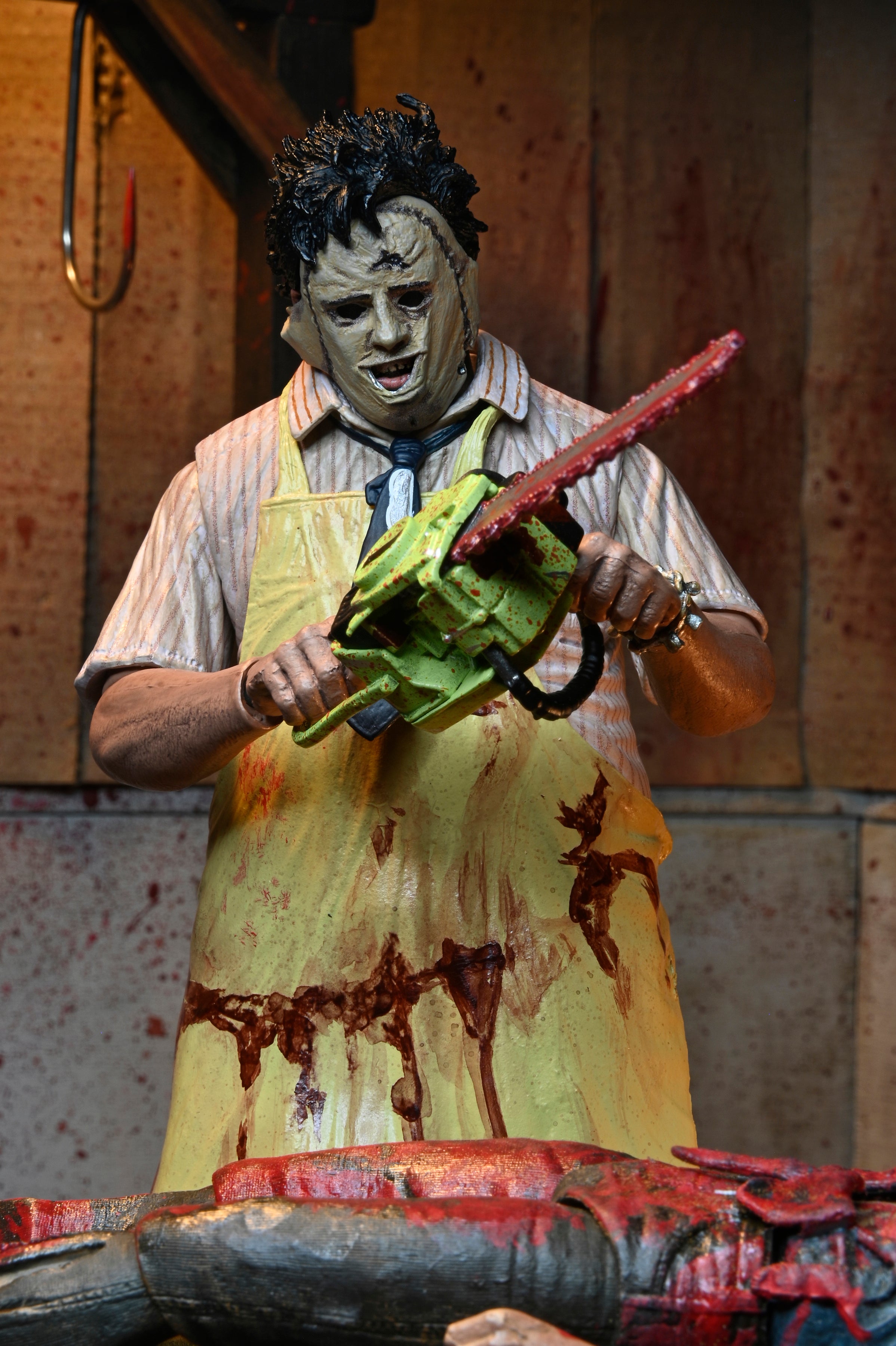 A figure clad in a grotesque mask and bloodied yellow apron, inspired by NECA’s Texas Chainsaw Massacre 50th Anniversary Leatherface Ultimate Action Figure (7 inch), menaces with a bloody chainsaw over a blood-soaked table in a dim, grim room.