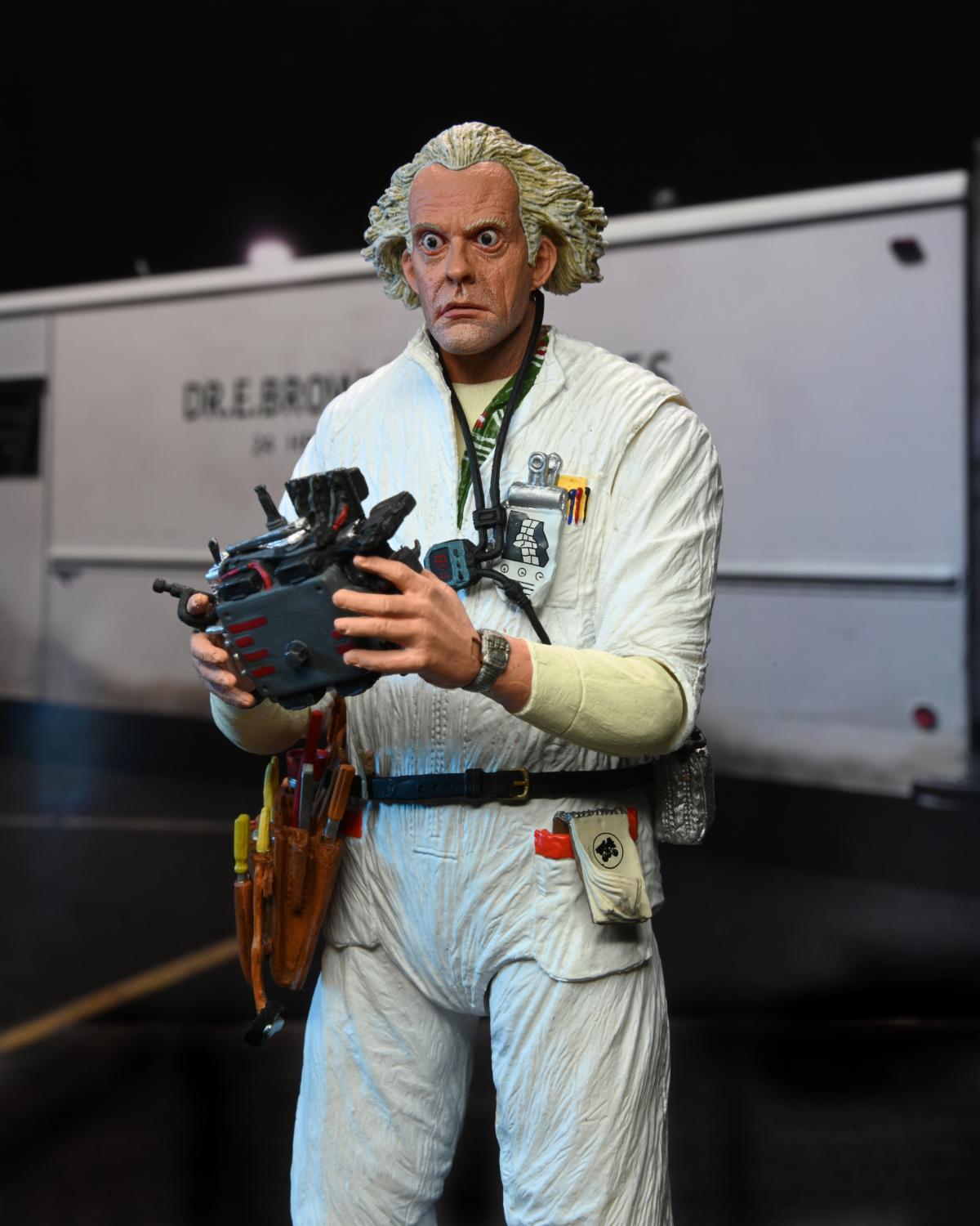 A wild white-haired figure in a white jumpsuit and tool belt holds a futuristic device before a DR. E. BROWN vehicle—just like the NECA Back to the Future Ultimate 1985 Doc Brown Hazmat Suit Action Figure (7 inch).