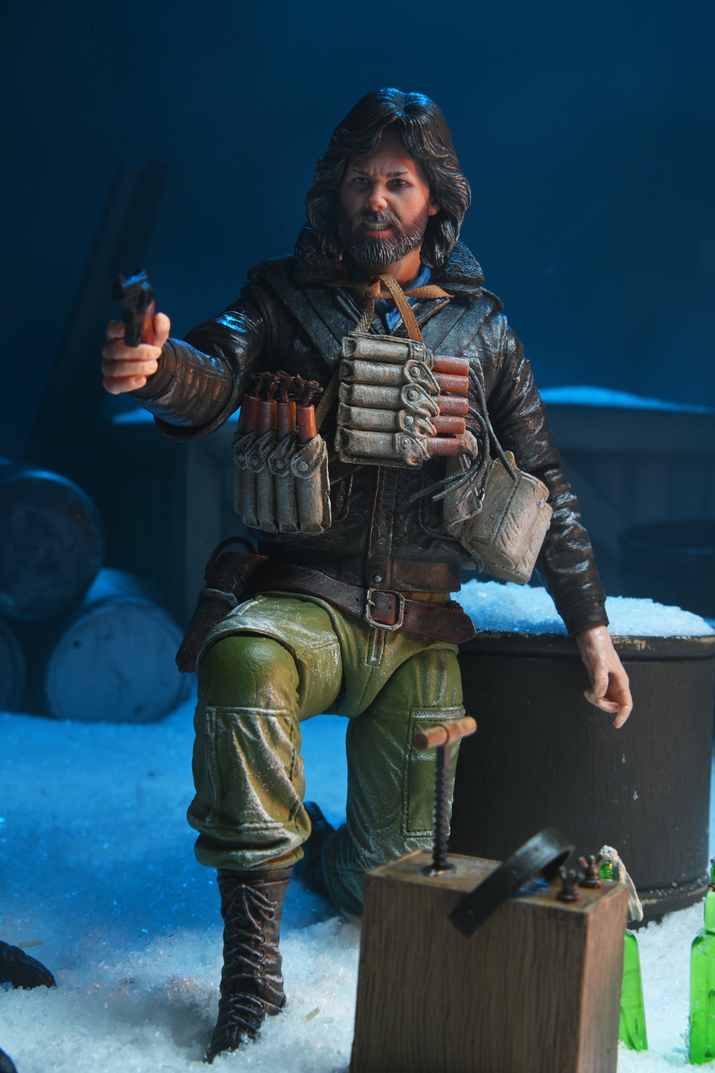 The NECA The Thing R.J. MacReady Last Stand Ultimate Action Figure (7 inch) depicts a bearded MacReady in winter gear, kneeling on snow with a pistol and dynamite bandolier, with barrels and supplies in the snowy background.