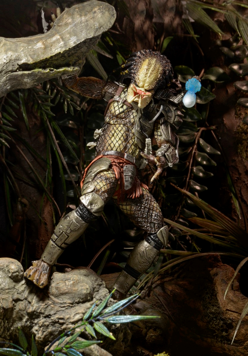 The NECA Predator Jungle Hunter Ultimate Action Figure (7 inch) stands in a jungle scene, dressed in armor and netted clothing, surrounded by foliage and rocks, holding a blue-tipped weapon.
