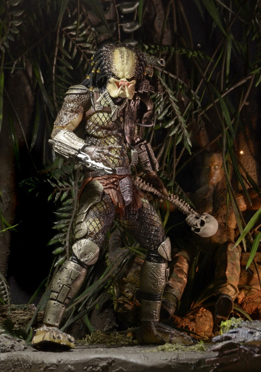 The NECA Predator Jungle Hunter Ultimate Action Figure (7 inch) stands in the jungle, holding a skull and spine. It features armor, a netted body suit, alien facial details, and dreadlock-like hair among lush foliage.