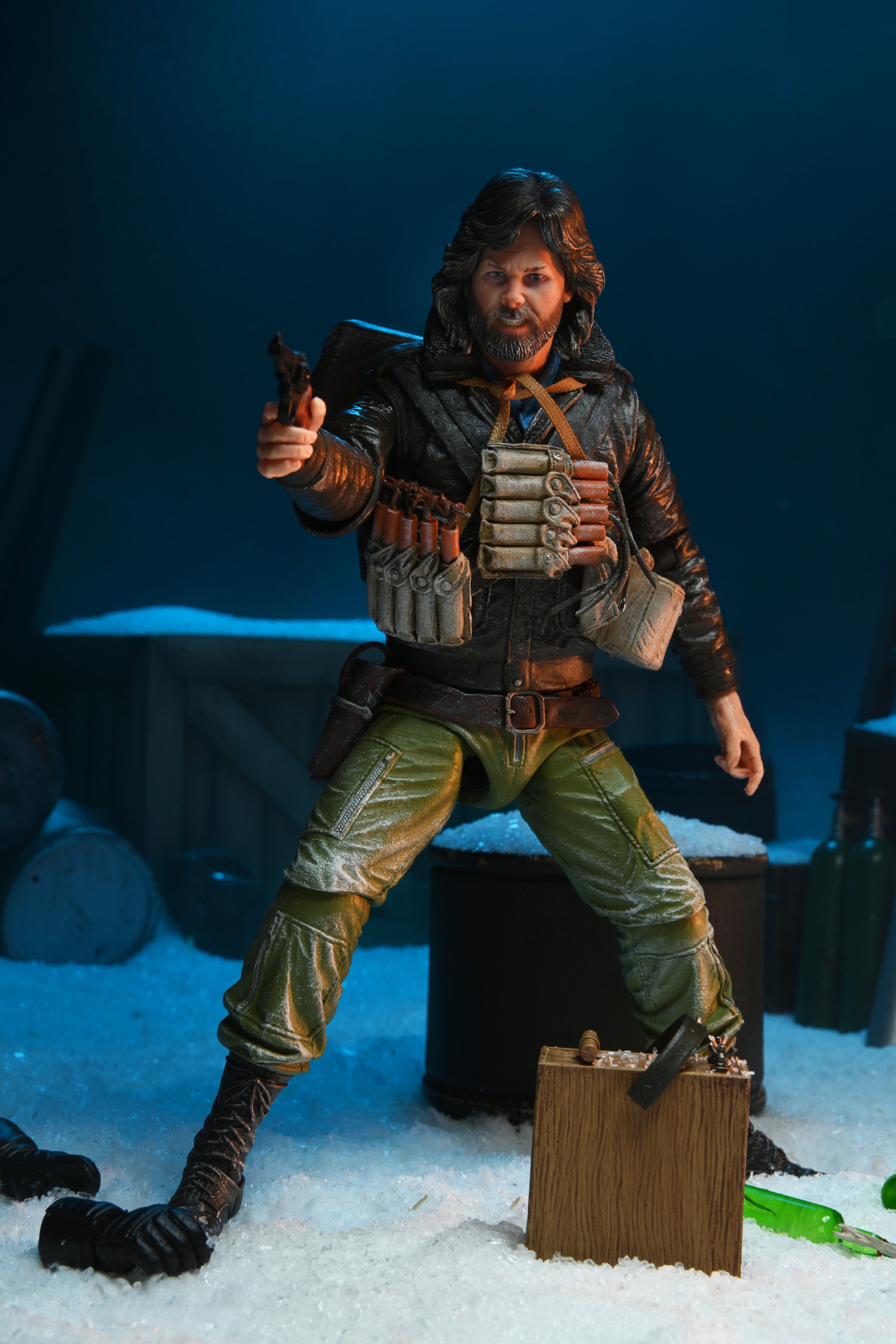 The NECA The Thing R.J. MacReady Last Stand Ultimate Action Figure (7 inch) features a bearded man in winter gear, aiming a gun with ammo on his chest and dynamite at his feet, standing in snowy scenery with crates and barrels.