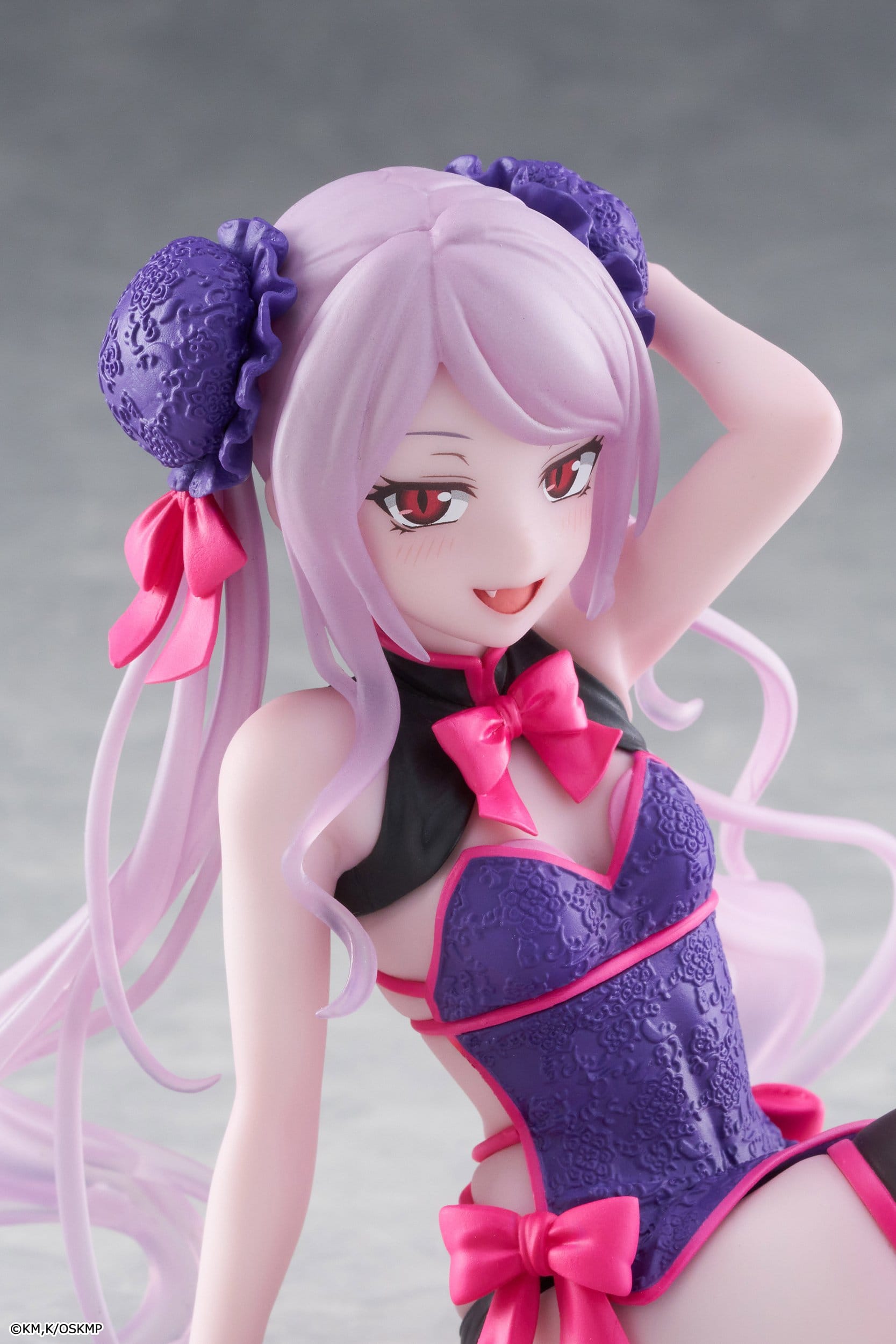 A close-up of the Taito Desktop Cute Shalltear Chinese Dress Ver. PVC Statue (13 cm) by Taito, featuring anime-style details: long light purple hair, red eyes, a playful smile, and a purple-pink outfit with bows on a plain gray background.